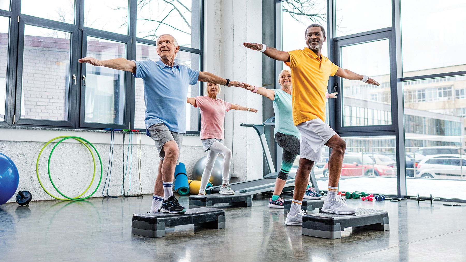 Group of seniors exercising in a bright gym, performing balance exercises on step platforms. The participants wear colorful workout clothes and smile as they follow the instructor's lead.