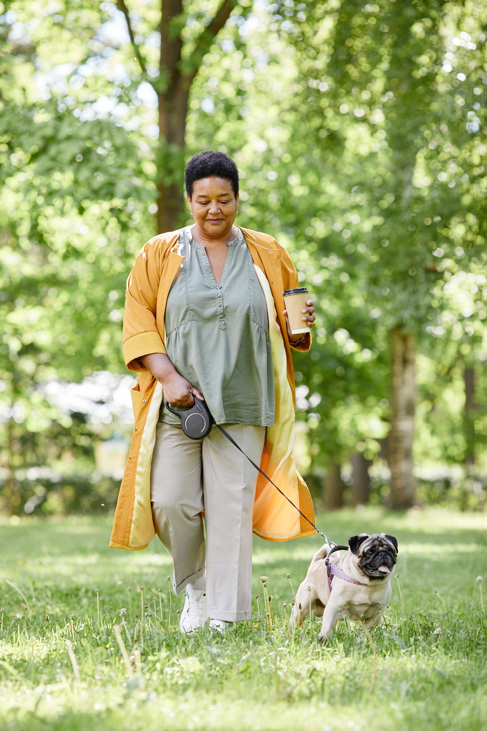 A woman in a yellow coat walks her dog in a park, holding a coffee cup and a leash, surrounded by lush greenery.