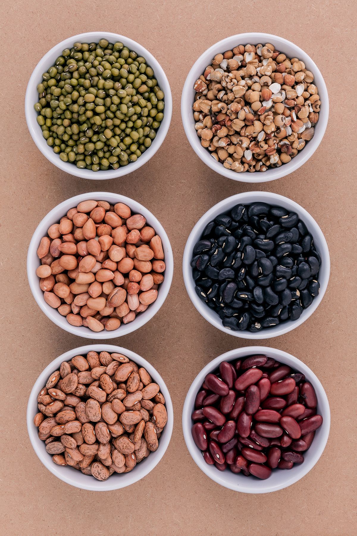 Six small white bowls filled with various dried beans and legumes, neatly arranged on a brown surface.