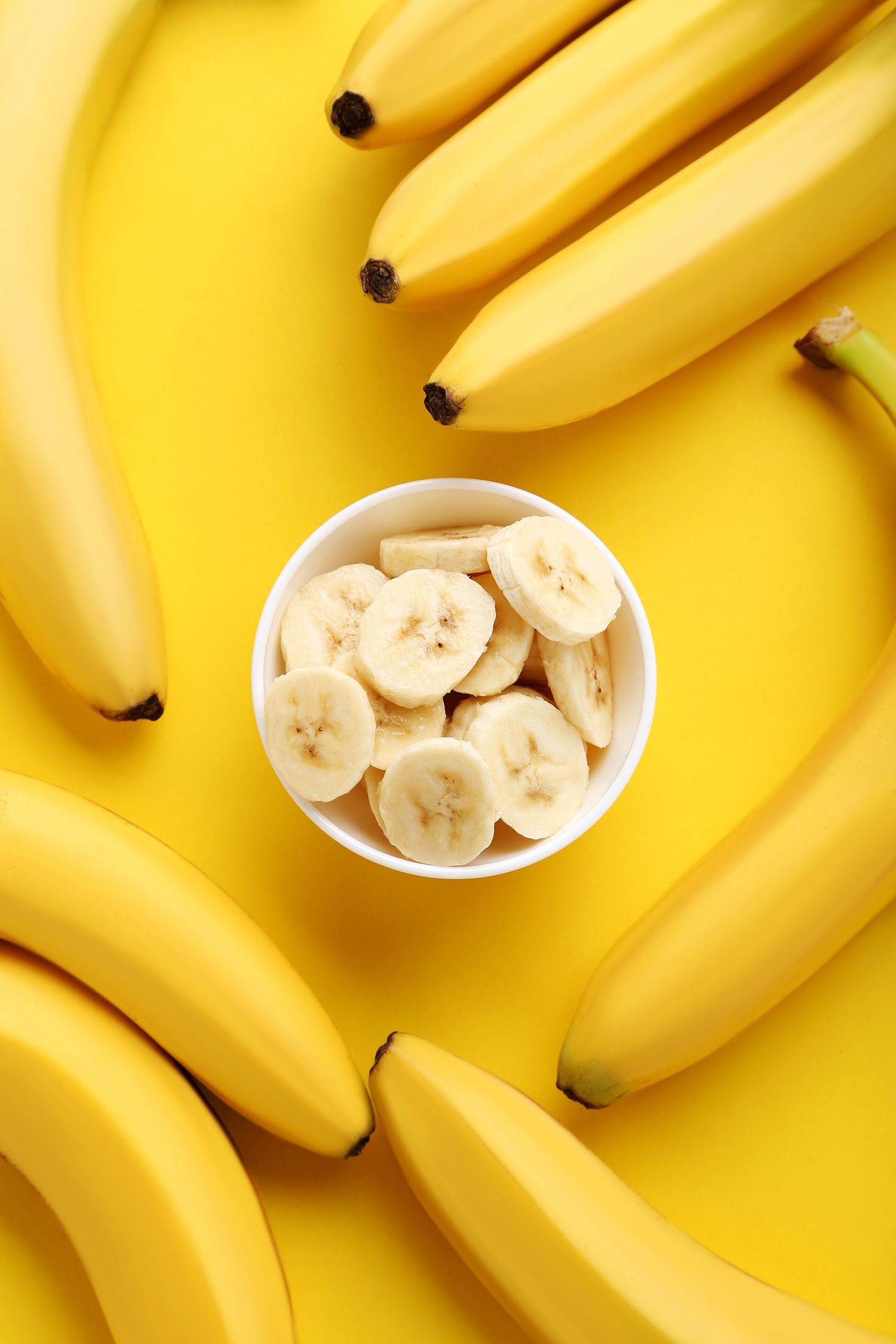 Overhead view of whole bananas surrounding a bowl of sliced bananas on a bright yellow background.