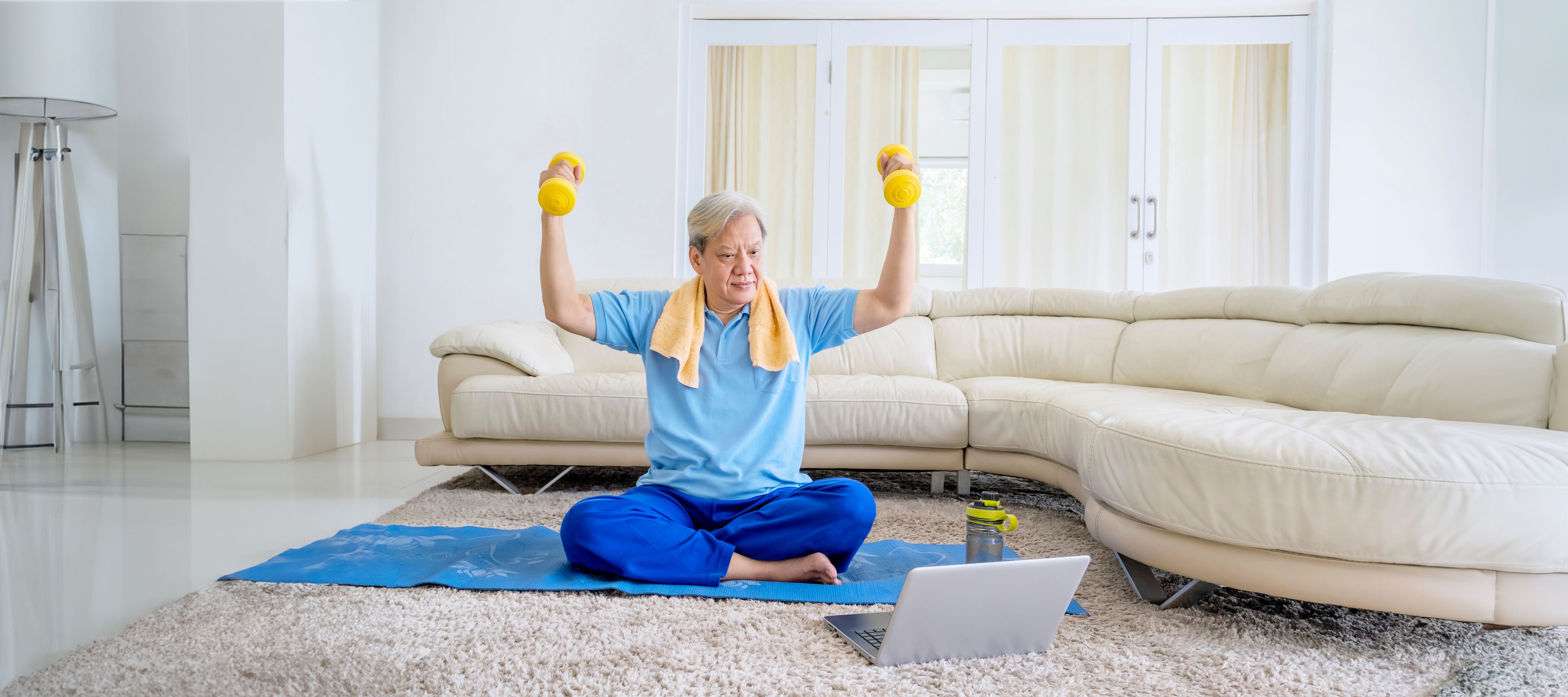 Senior man in a blue shirt and sweatpants lifting dumbbells while sitting on a yoga mat in his living room, participating in an online workout class.