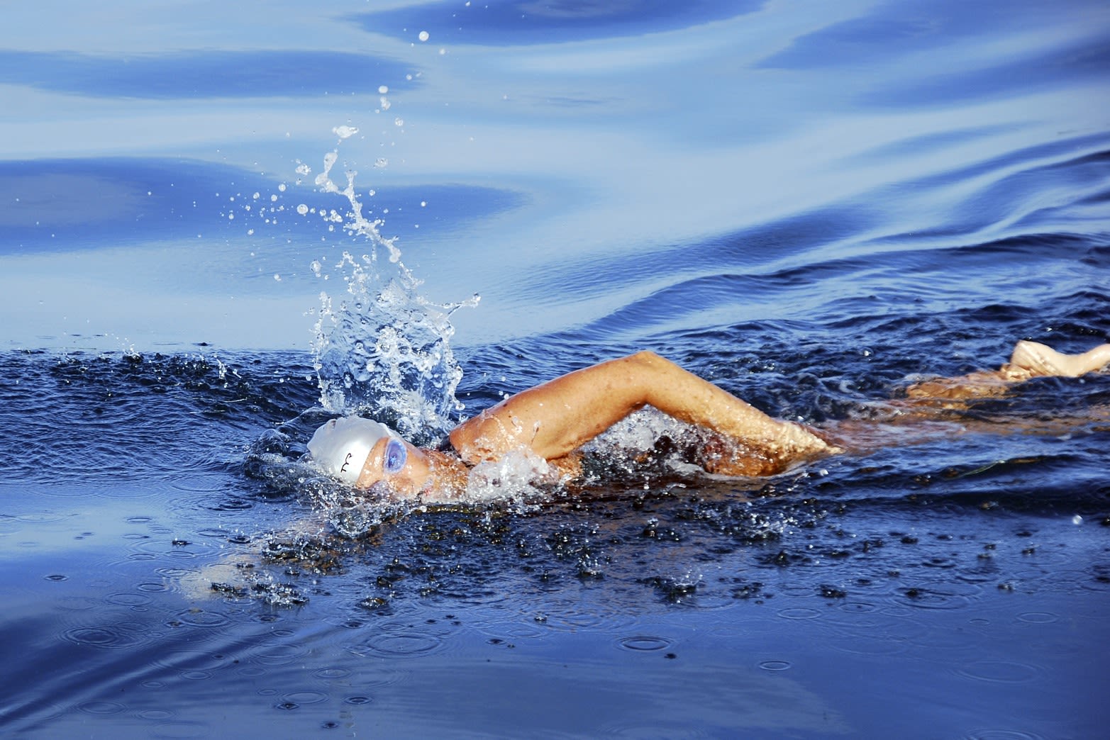 Diana Nyad wearing a white swim cap and goggles is swimming in open water, creating splashes as she moves through the blue water.