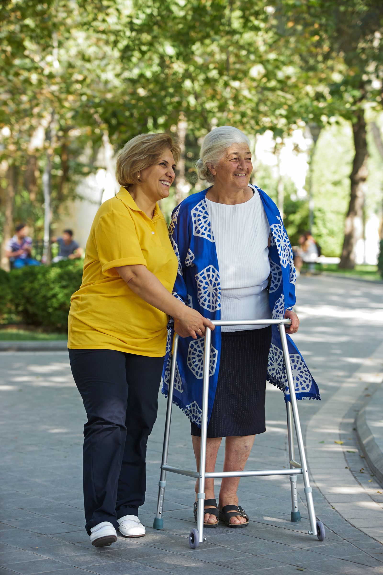 A cheerful caregiver in a yellow shirt walks alongside an elderly woman using a walker in a park. Both women are smiling and enjoying their time outdoors.