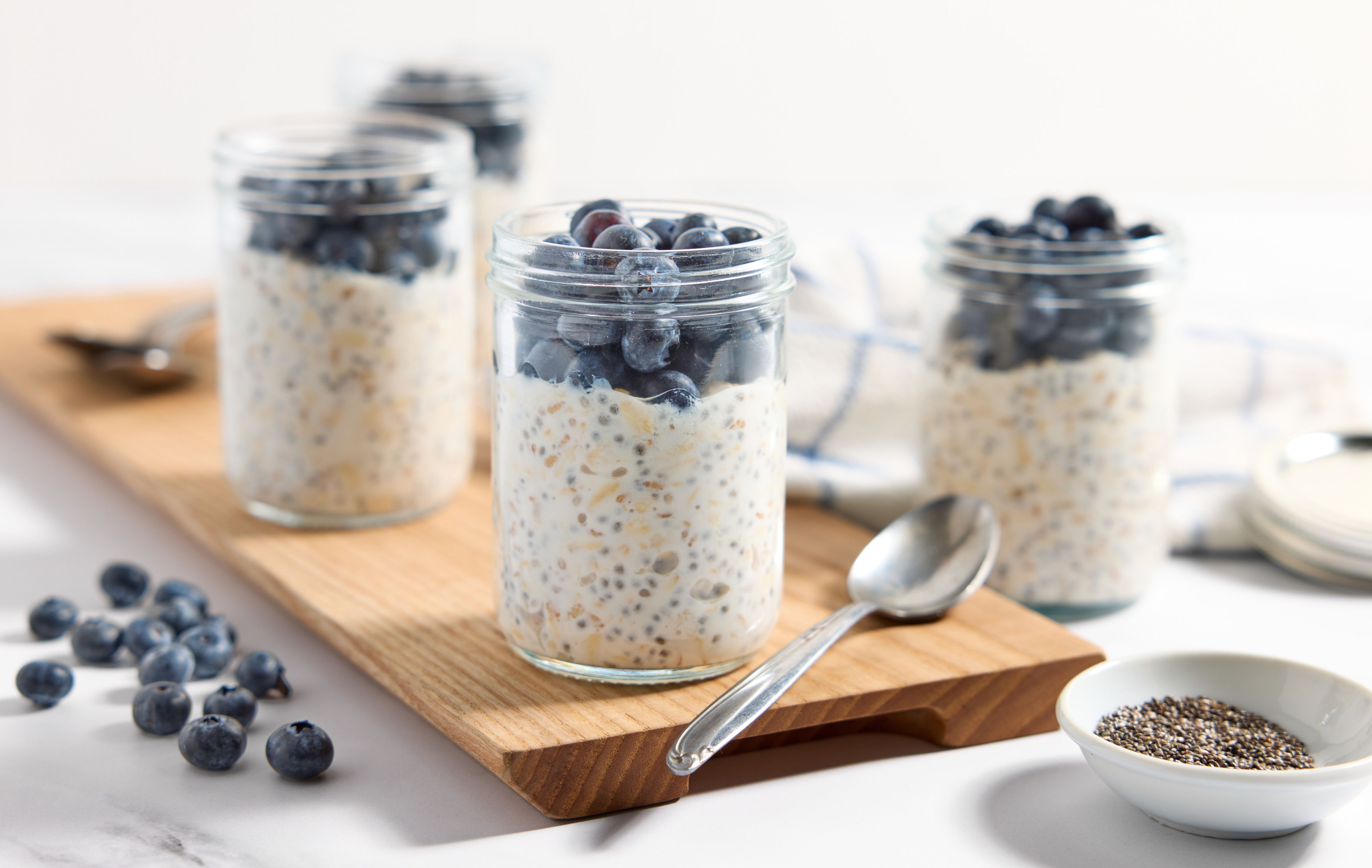 Four jars of blueberry overnight oats sit on a wooden board with spoons, a small bowl of chia seeds, and blueberries strewn to the side.