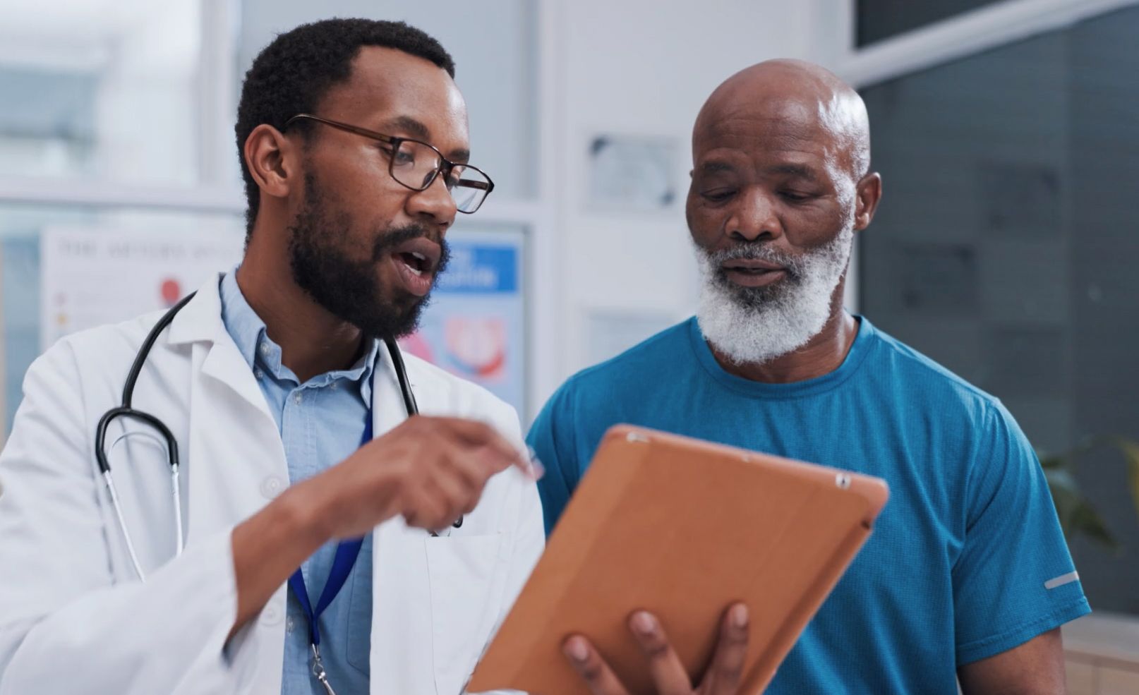 A doctor in a white coat and stethoscope is explaining something on a tablet to an older patient with a white beard and wearing a blue shirt.