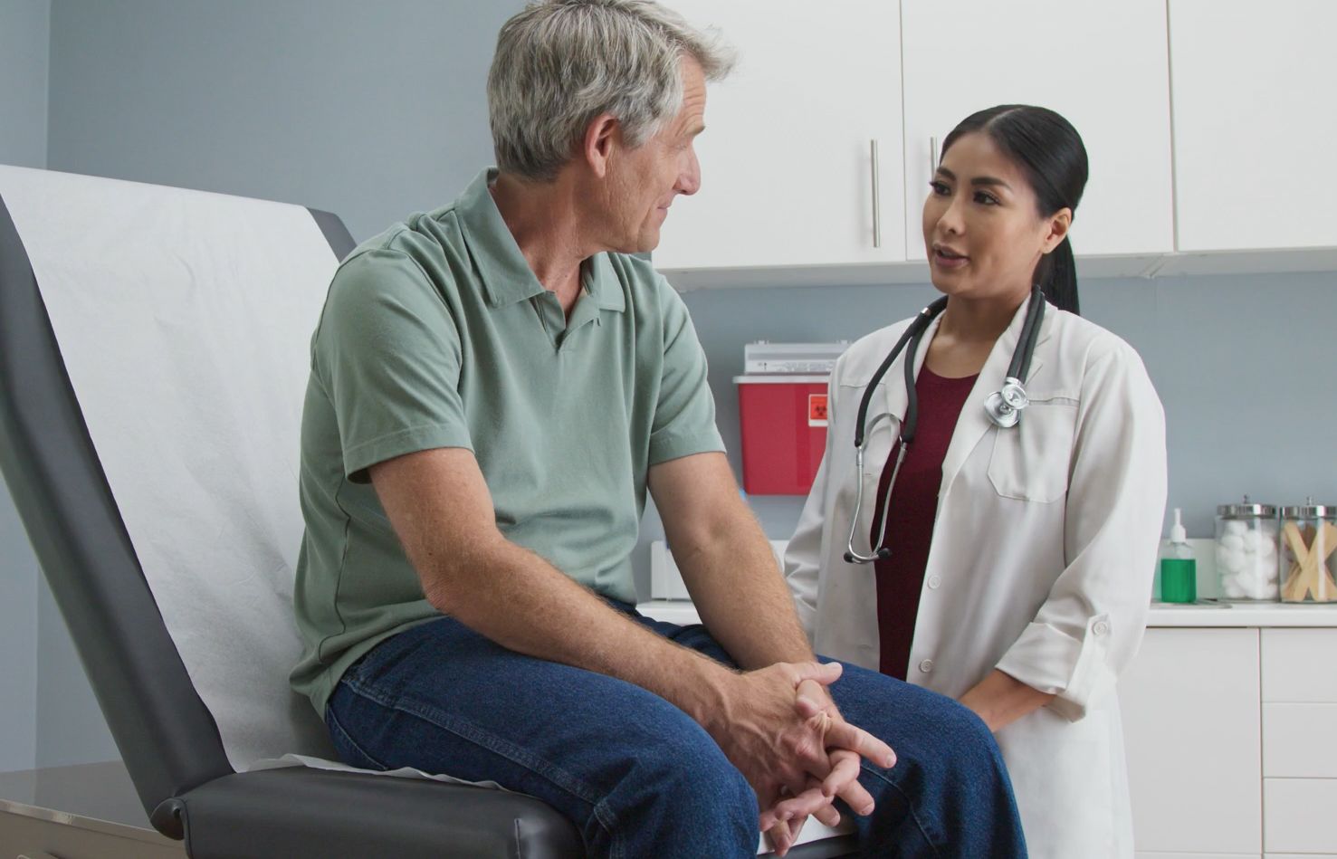 A female doctor in a white coat talks with a seated male patient in a green polo shirt, inside a medical examination room.