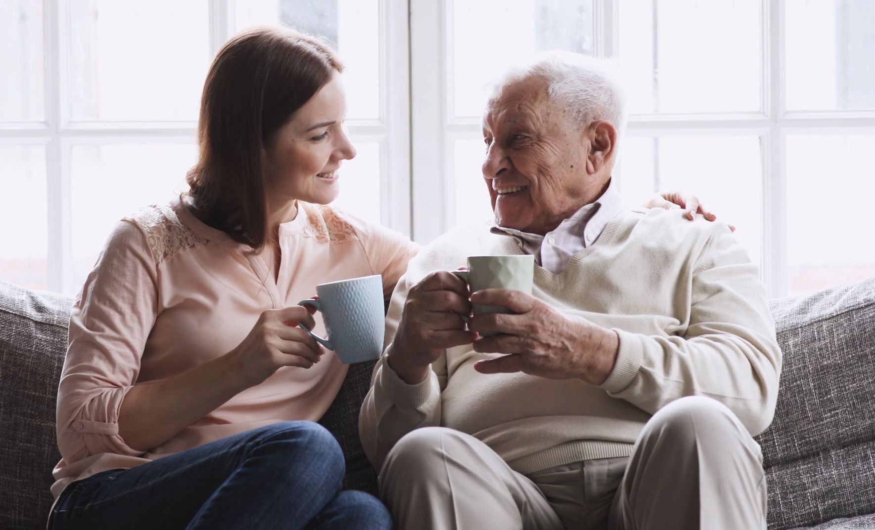 A smiling young woman and an elderly man enjoy a conversation while holding mugs of coffee or tea, seated on a couch in a bright, cozy room.