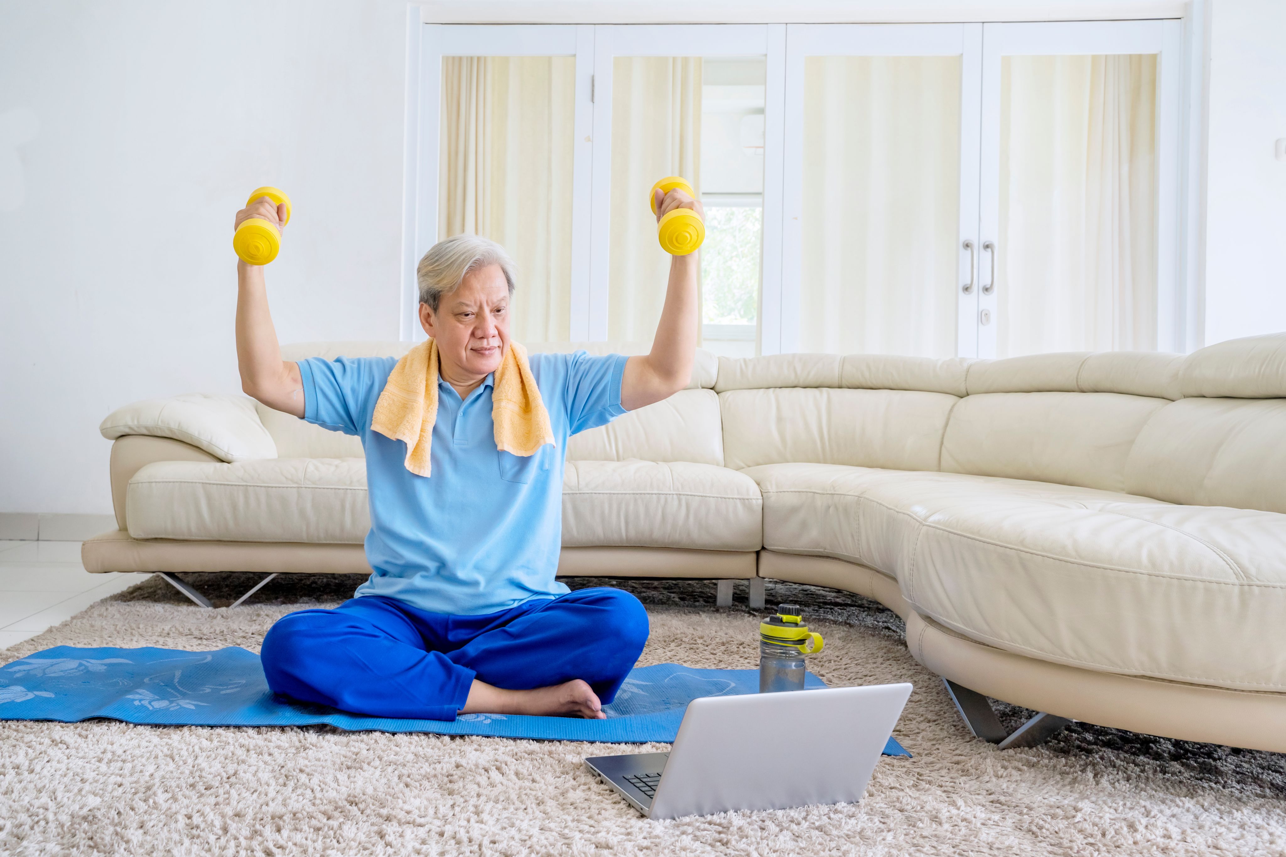 Senior man in a blue shirt and sweatpants lifting dumbbells while sitting on a yoga mat in his living room, participating in an online workout class.