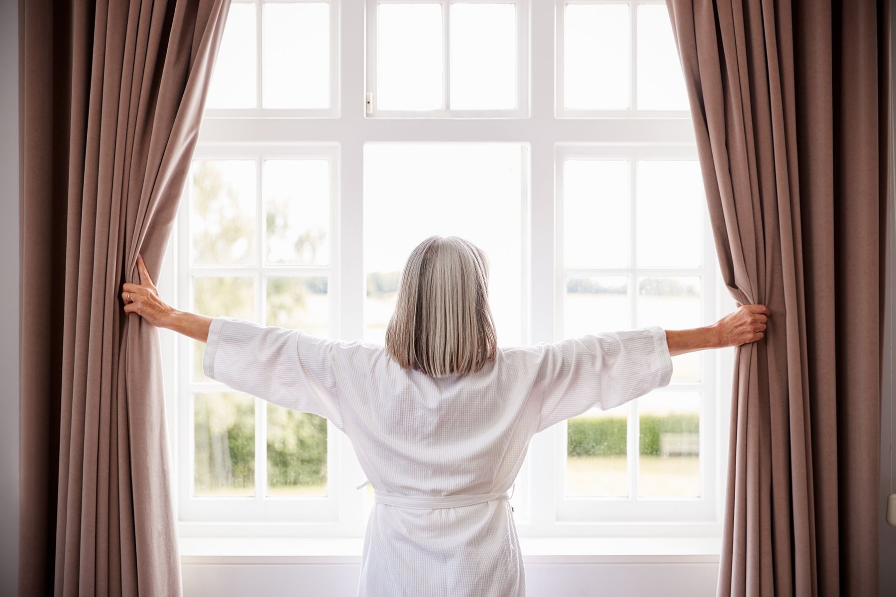 An older woman with gray hair, seen from behind, opens beige curtains to a bright morning outside. She is wearing a white bathrobe and stands in front of a large window, welcoming the sunlight into the room.
