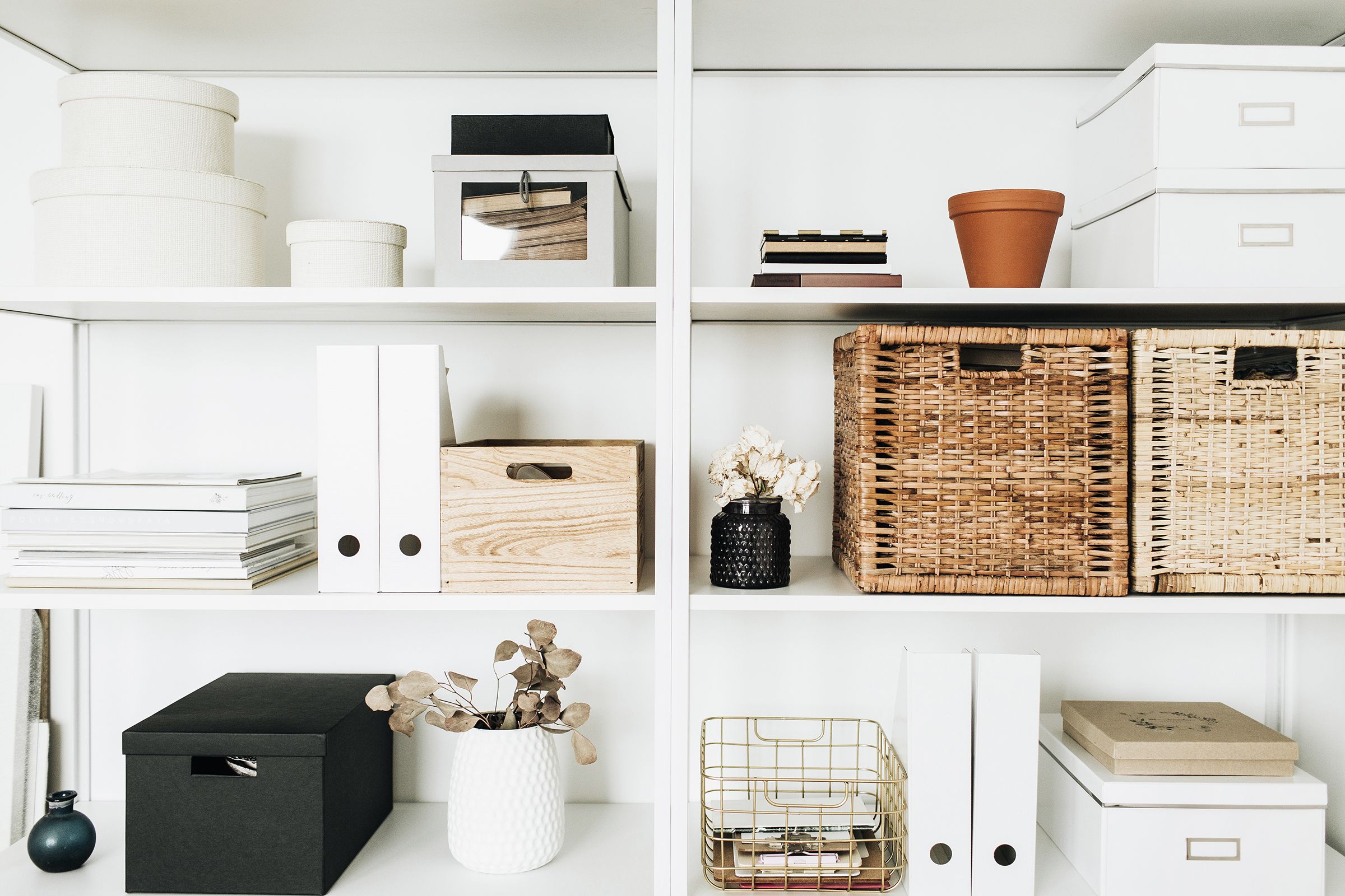 Neatly organized white shelves with various storage items, including woven baskets, white binders, decorative boxes, books, a terracotta pot, and vases with dried flowers, creating a clean and minimalist aesthetic.