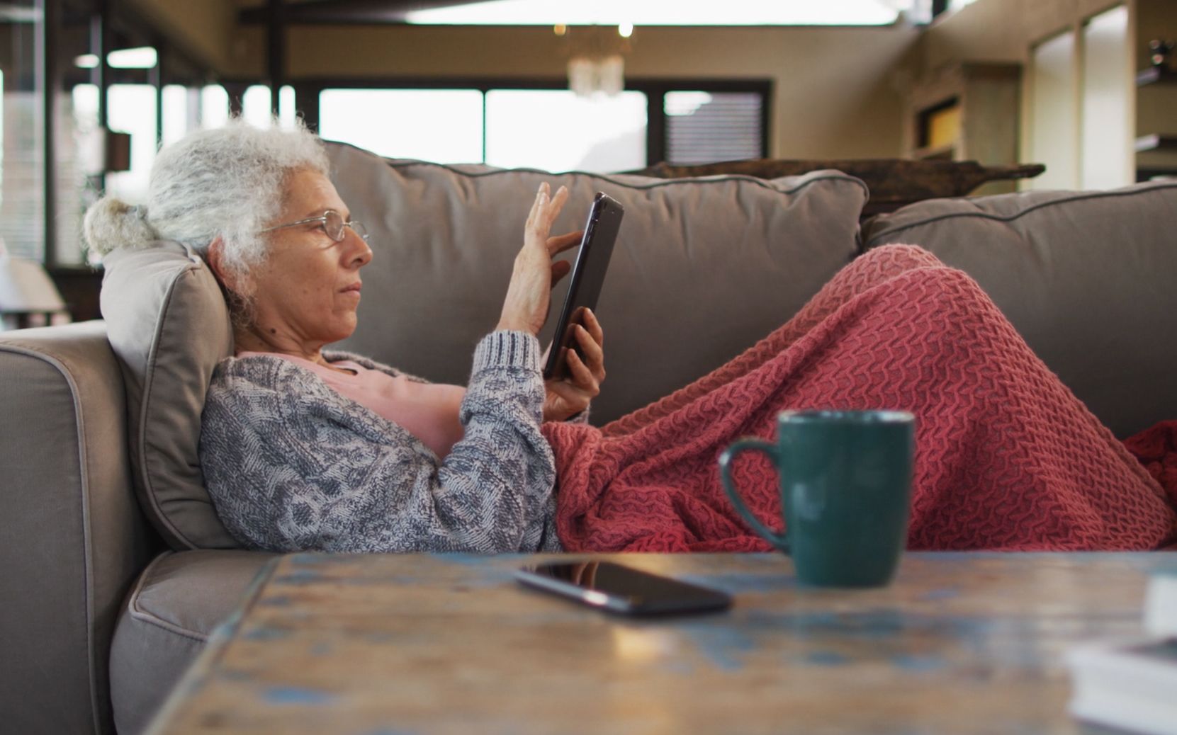 An older woman with gray hair and glasses, wearing a gray sweater and pink shirt, is sitting on a couch under a red blanket, using a tablet. A green mug and a smartphone are on the table in front of her.