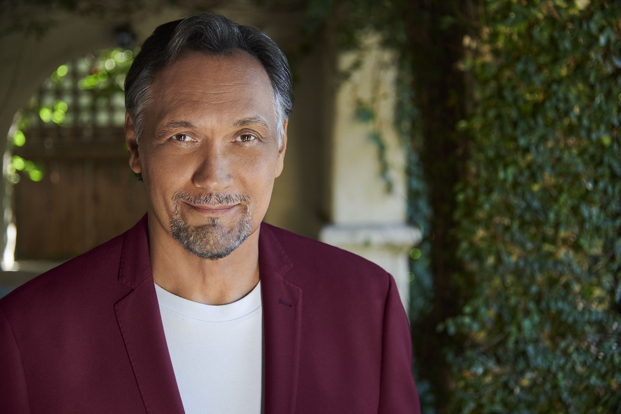 Image of actor Jimmy Smits in a red suit jacket and white t-shirt in a courtyard looking at the camera.