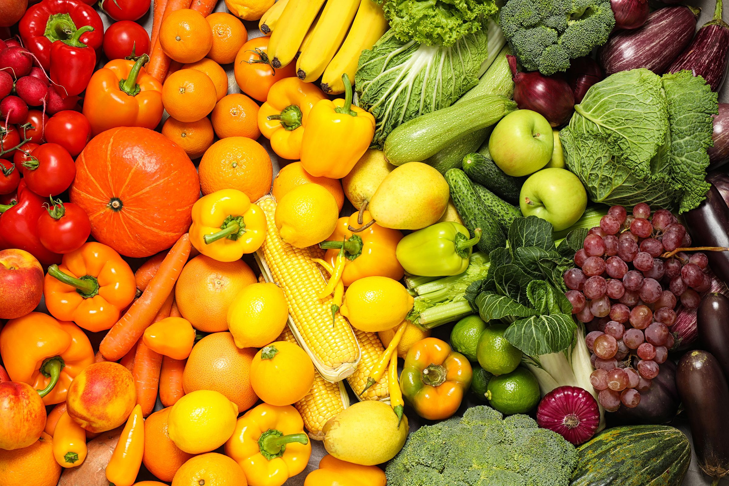 Assorted fresh fruits and vegetables arranged in a colorful display, featuring red tomatoes, radishes, and bell peppers, orange pumpkins, carrots, and oranges, yellow bananas and corn, green lettuce, cucumbers, and apples, purple grapes, and eggplants.